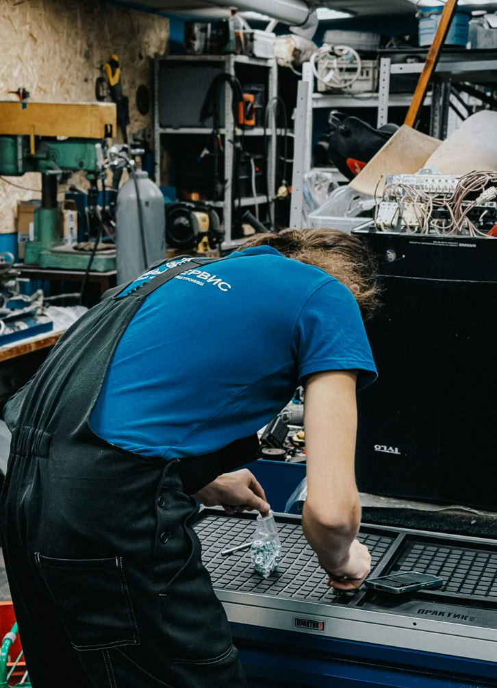A technician repairs equipment in a cluttered workshop setting. Industrial tools surround them.